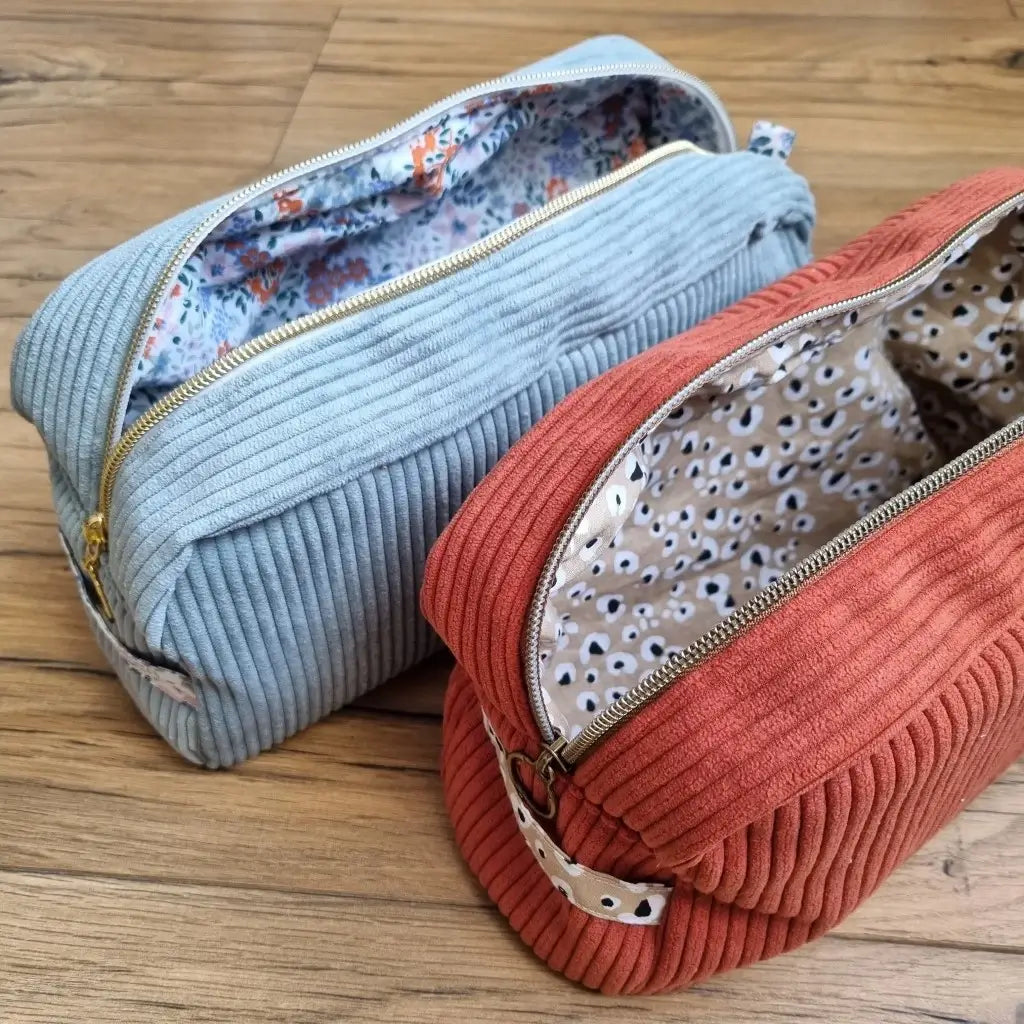 Deux trousses de toilettes en velours côtelé disposées côte à côte au sol sur un parquet en chêne. Au premier plan, la trousse de toilette en velours côtelé de couleur terracotta est ouverte et laisse apparaître un tissu intérieur au motif léopard. Au second plan, la trousse de toilette en velours côtelé de couleur bleu ciel est également ouverte laissant apparaître un tissu intérieur au motif fleuri.