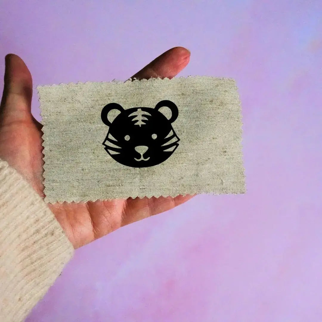 Main tenant une étiquette en tissu beige avec un motif de tête de tigre noir minimaliste, sur fond lavande.