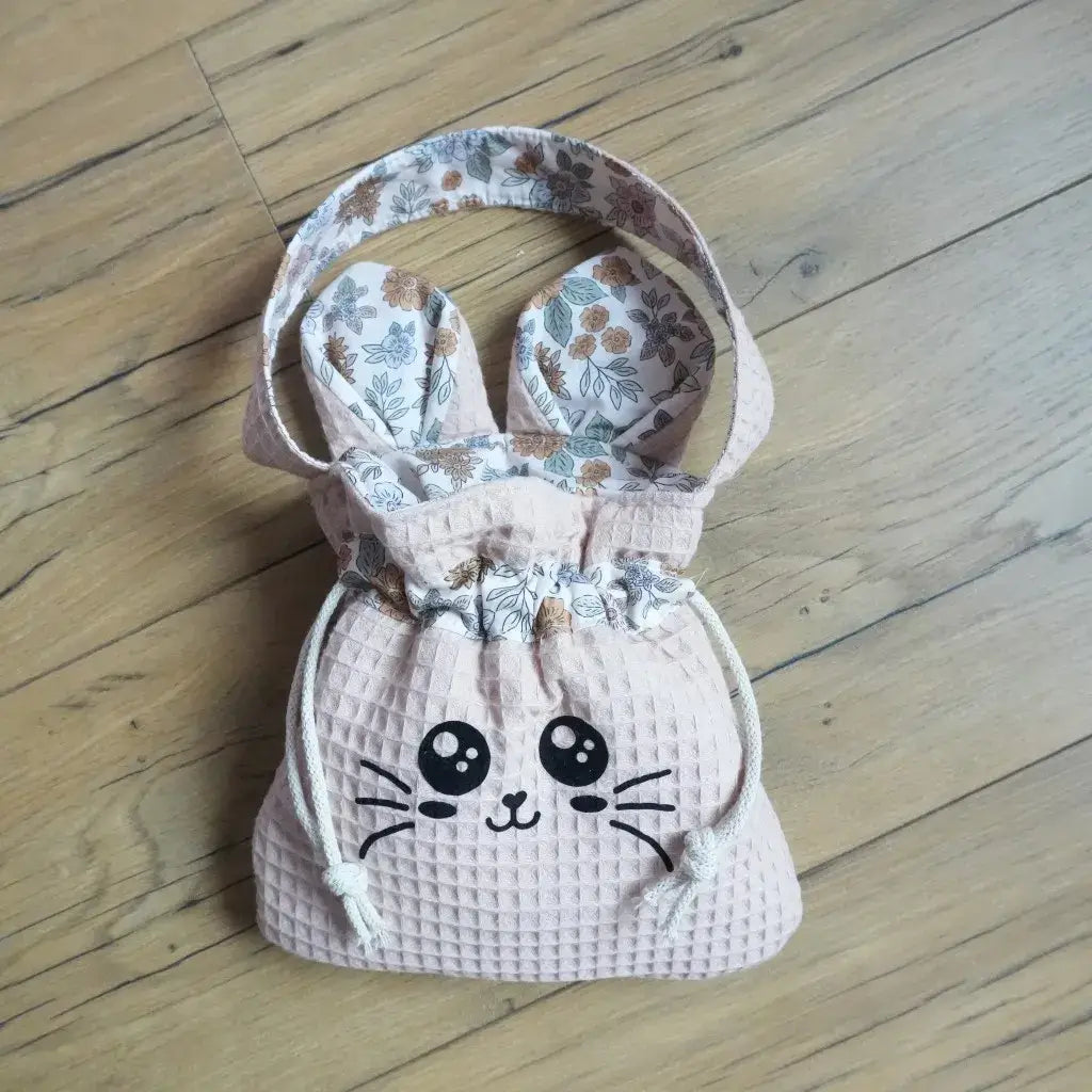 Pochon rose en nid d'abeille avec flocage chat noir et oreilles fleuries, posé sur un parquet en bois clair.