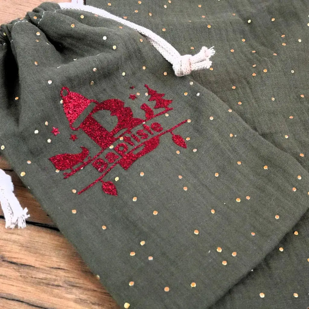 Flocage personnalisé en flex thermocollant rouge pailleté avec initiale et prénom Baptiste sur pochon vert en double gaze. Motif de Noël scintillant avec bonnet, sapin et décor étoilé, parfait pour sublimer un emballage cadeau ou accessoire cousu main. Personnalisation textile durable et festive pour créations couture uniques.