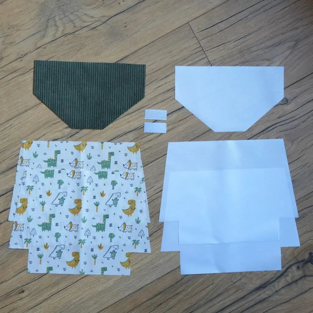 Kit couture DIY pour réaliser une pochette à scratch imperméable, comprenant un rabat en velours côtelé vert, des coupons de coton blanc imprimé de motifs dinosaures colorés, des doublures blanches en coton enduit et deux bandes de scratch. Ce kit prêt-à-coudre permet de créer une trousse originale et ludique, parfaite pour les enfants et idéale comme projet créatif ou cadeau fait main personnalisé.