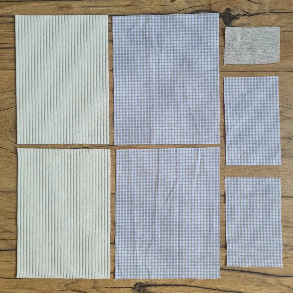 Kit "prêt à coudre" pour une pochette à livre & liseuse en velours côtelé au coloris beige avec une doublure en tissu à motif carreaux beiges. L'ensemble des pièces de tissu, déjà prédécoupées aux bonnes dimensions, sont disposées au sol. A gauche, deux pièces identiques en velours côtelé beige. A droite, quatre pièces en tissu imprimé à motif carreaux beiges ainsi qu’une étiquette en tissu toile uni servant de support à une inscription personnalisée qui sera floquée dessus.