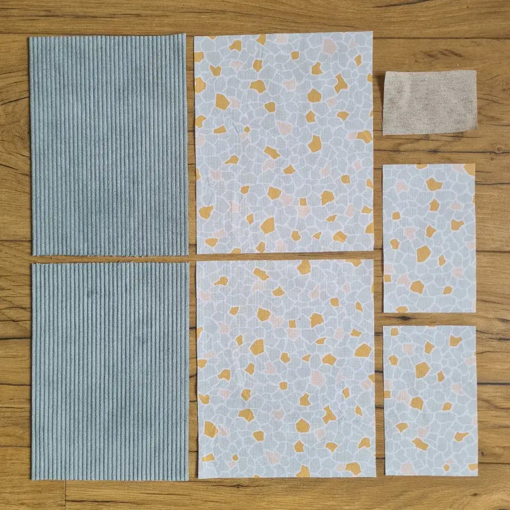 Kit "prêt à coudre" pour une pochette à livre & liseuse en velours côtelé au coloris bleu clair avec une doublure en tissu à motif argeo. L'ensemble des pièces de tissu, déjà prédécoupées aux bonnes dimensions, sont disposées au sol. A gauche, deux pièces identiques en velours côtelé bleu clair. A droite, quatre pièces en tissu imprimé à motif argeo ainsi qu’une étiquette en tissu toile uni servant de support à une inscription personnalisée qui sera floquée dessus.