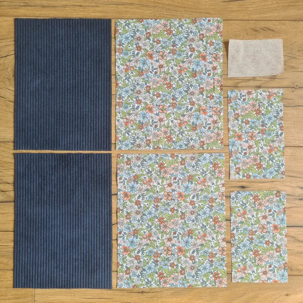 Kit "prêt à coudre" pour une pochette à livre & liseuse en velours côtelé au coloris bleu foncé avec une doublure en tissu à motif fleuri. L'ensemble des pièces de tissu, déjà prédécoupées aux bonnes dimensions, sont disposées au sol. A gauche, deux pièces identiques en velours côtelé bleu foncé. A droite, quatre pièces en tissu imprimé à motif fleuri ainsi qu’une étiquette en tissu toile uni servant de support à une inscription personnalisée qui sera floquée dessus.