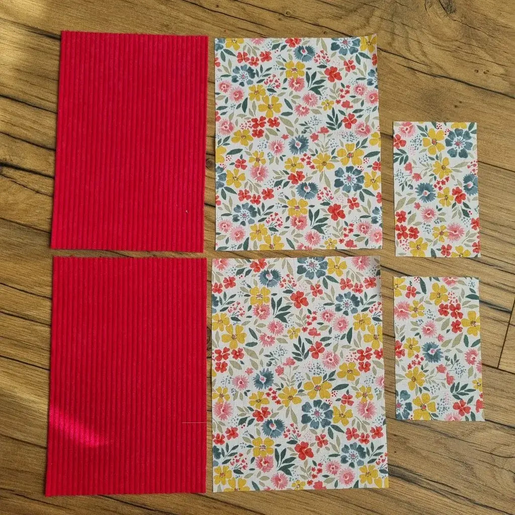 Kit prêt-à-coudre composé de pièces de tissu découpées, prêtes à assembler pour réaliser une pochette à livre ou liseuse. Le lot comprend du velours côtelé uni couleur framboise et un tissu imprimé floral multicolore aux tons jaune, rouge, bleu et vert, disposés à plat sur une table en bois. Chaque élément est soigneusement préparé pour faciliter la création d’un accessoire unique et fait main.