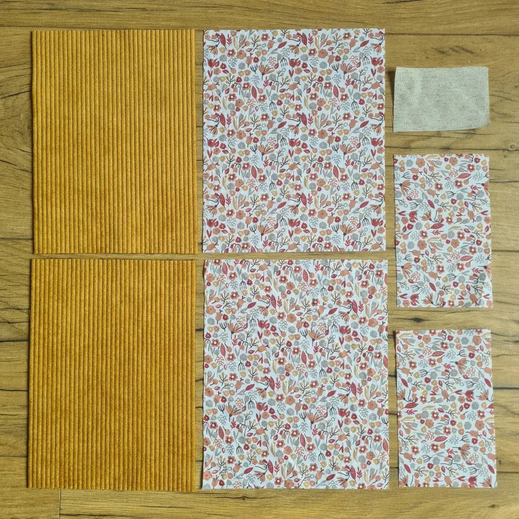 Kit "prêt à coudre" pour une pochette à livre & liseuse en velours côtelé au coloris jaune avec une doublure en tissu à motif liberty. L'ensemble des pièces de tissu, déjà prédécoupées aux bonnes dimensions, sont disposées au sol. A gauche, deux pièces identiques en velours côtelé jaune. A droite, quatre pièces en tissu imprimé à motif liberty ainsi qu’une étiquette en tissu toile uni servant de support à une inscription personnalisée qui sera floquée dessus.
