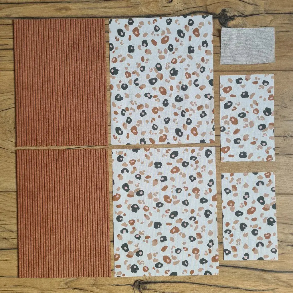 Kit "prêt à coudre" pour une pochette à livre & liseuse en velours côtelé au coloris marron avec une doublure en tissu à motif léopard. L'ensemble des pièces de tissu, déjà prédécoupées aux bonnes dimensions, sont disposées au sol. A gauche, deux pièces identiques en velours côtelé marron. A droite, quatre pièces en tissu imprimé à motif léopard ainsi qu’une étiquette en tissu toile uni servant de support à une inscription personnalisée qui sera floquée dessus.