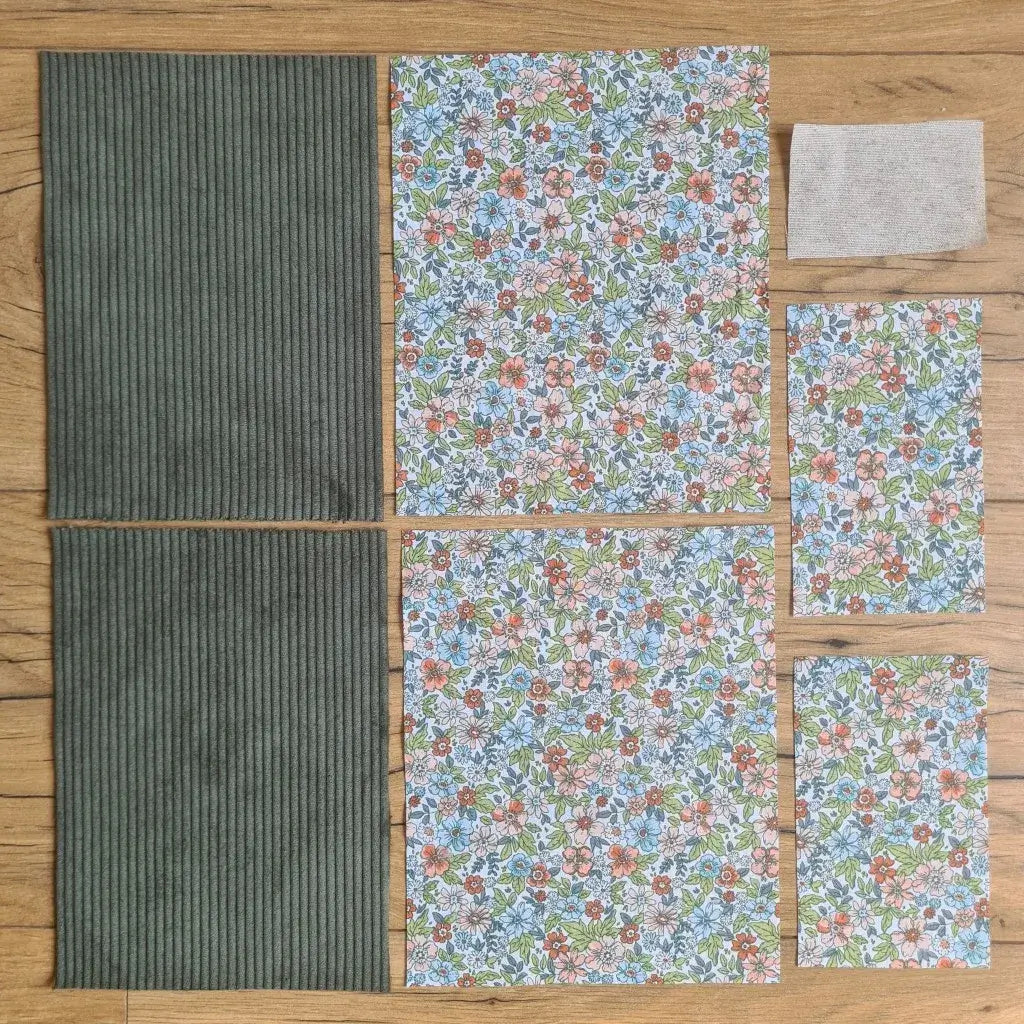 Kit "prêt à coudre" pour une pochette à livre & liseuse en velours côtelé au coloris vert avec une doublure en tissu à motif fleuri. L'ensemble des pièces de tissu, déjà prédécoupées aux bonnes dimensions, sont disposées au sol. A gauche, deux pièces identiques en velours côtelé vert. A droite, quatre pièces en tissu imprimé à motif fleuri ainsi qu’une étiquette en tissu toile uni servant de support à une inscription personnalisée qui sera floquée dessus.