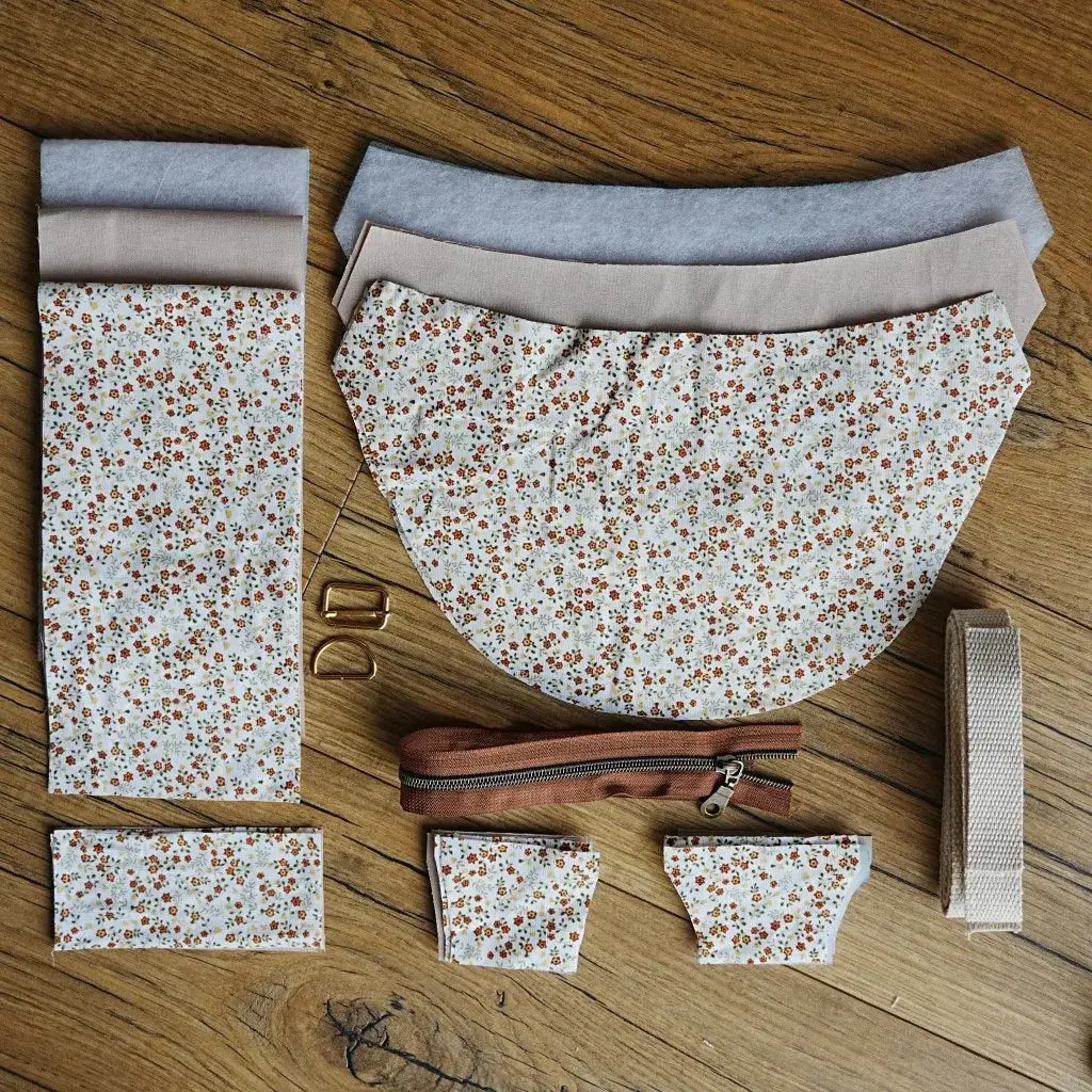 Kit de couture pour sac demi-lune comprenant des pièces de tissu coton imprimé fleuri, doublure unie coloris lin, renfort molletonné, sangle en coton écru, fermeture éclair marron et boucles métalliques dorées, disposés sur un sol en bois. Ensemble complet idéal pour réaliser un sac tendance et personnalisé, parfait pour les projets DIY couture.
