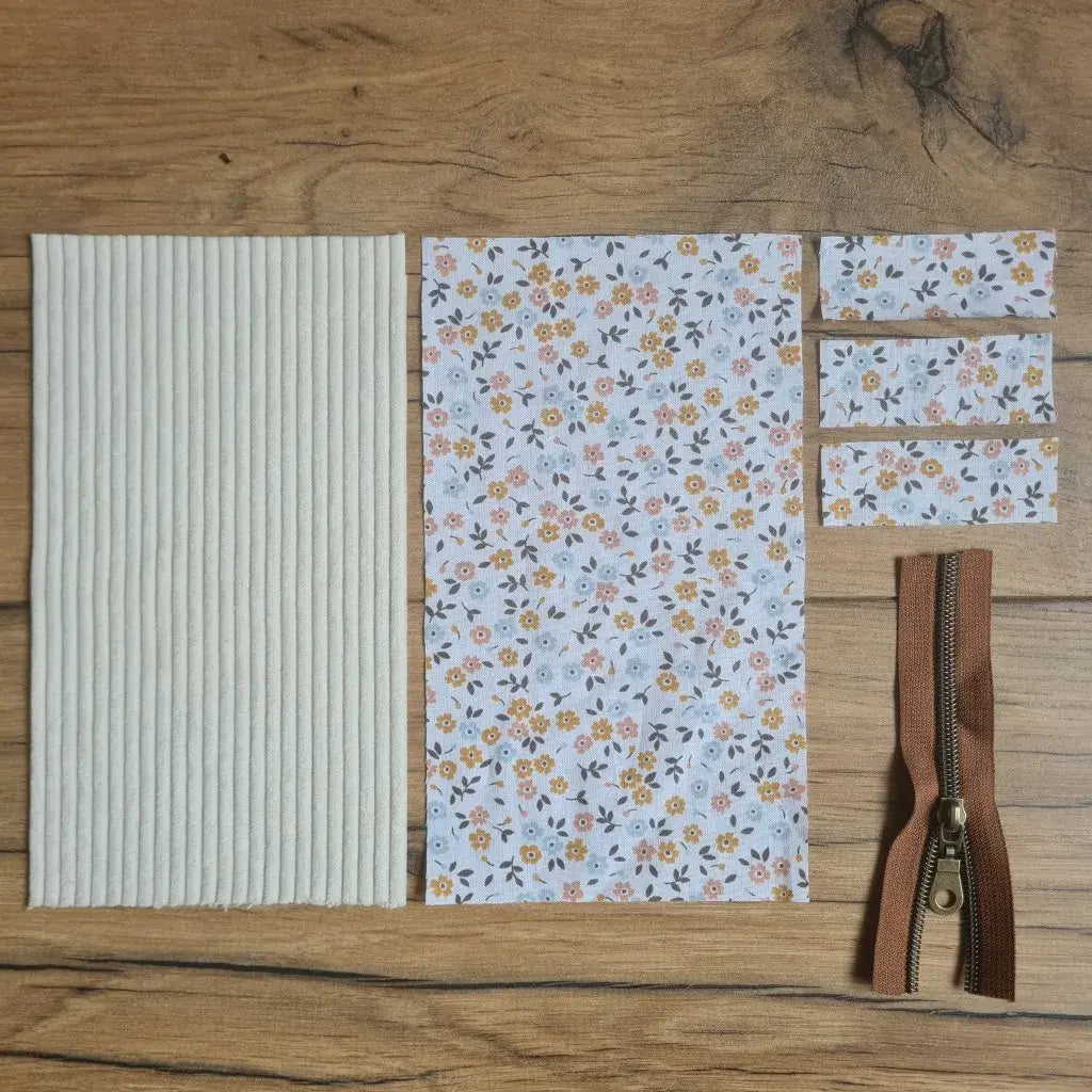 Kit "prêt à coudre" pour une trousse berlingot en velours côtelé au coloris beige avec une doublure en tissu à motif fleurette. L'ensemble des pièces de tissu, déjà prédécoupées aux bonnes dimensions, sont disposées au sol. A gauche, une pièce en velours côtelé beige. A droite, quatre pièces en tissu imprimé à motif fleurette ainsi qu'une fermeture éclair marron.