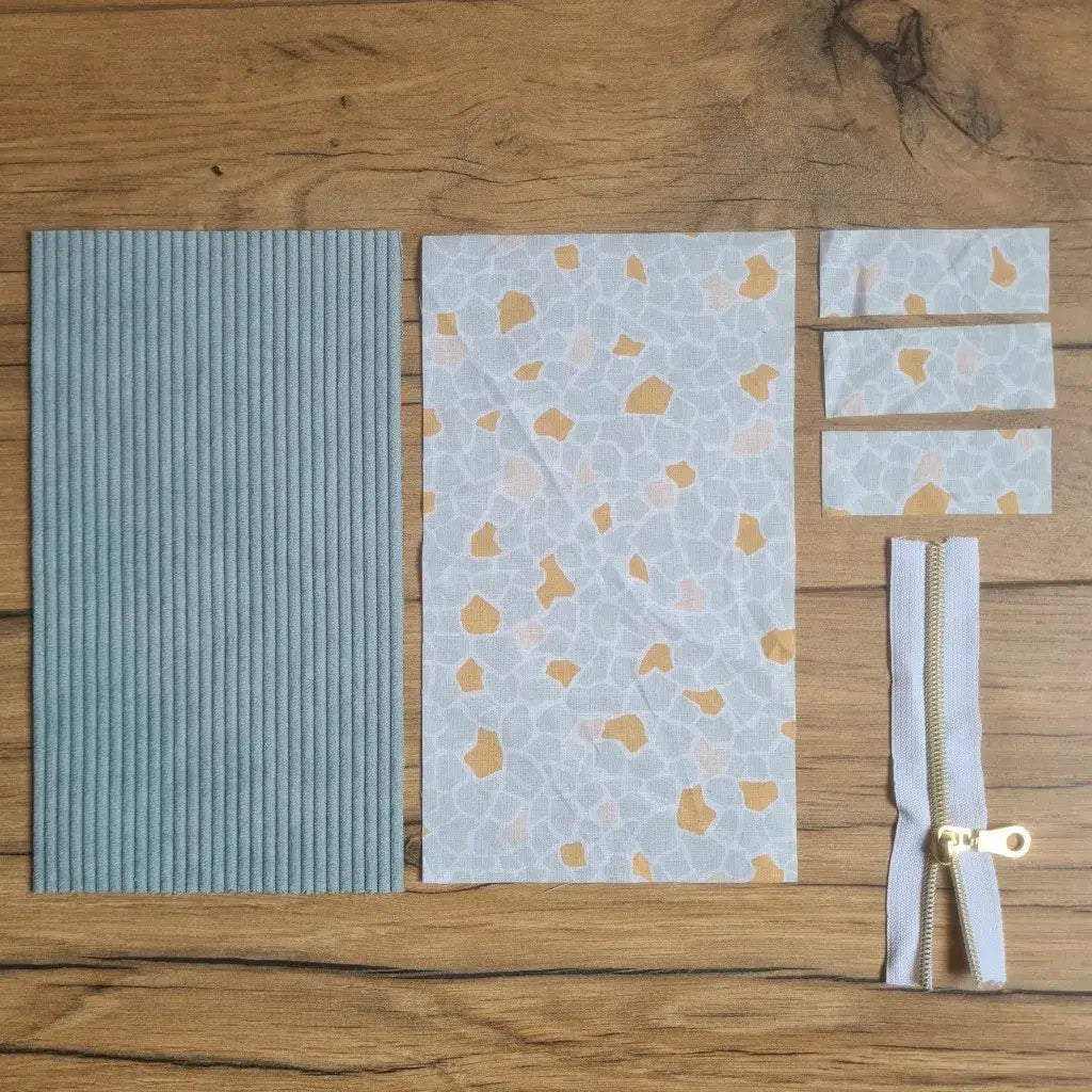 Kit "prêt à coudre" pour une trousse berlingot en velours côtelé au coloris bleu clair avec une doublure en tissu à motif argeo. L'ensemble des pièces de tissu, déjà prédécoupées aux bonnes dimensions, sont disposées au sol. A gauche, une pièce en velours côtelé bleu clair. A droite, quatre pièces en tissu imprimé à motif argeo ainsi qu'une fermeture éclair blanche.