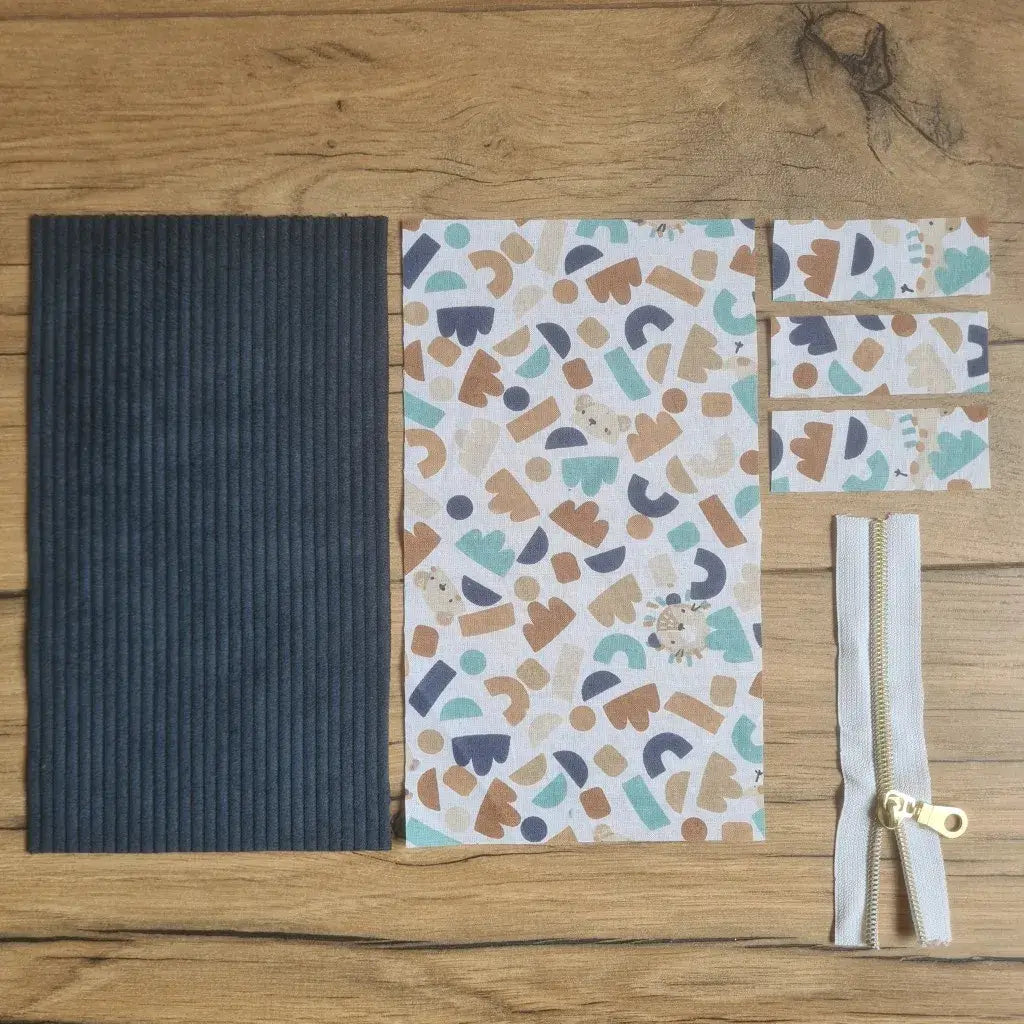 Kit "prêt à coudre" pour une trousse berlingot en velours côtelé au coloris bleu foncé avec une doublure en tissu à motif ptiwi. L'ensemble des pièces de tissu, déjà prédécoupées aux bonnes dimensions, sont disposées au sol. A gauche, une pièce en velours côtelé bleu foncé. A droite, quatre pièces en tissu imprimé à motif ptiwi ainsi qu'une fermeture éclair blanche.
