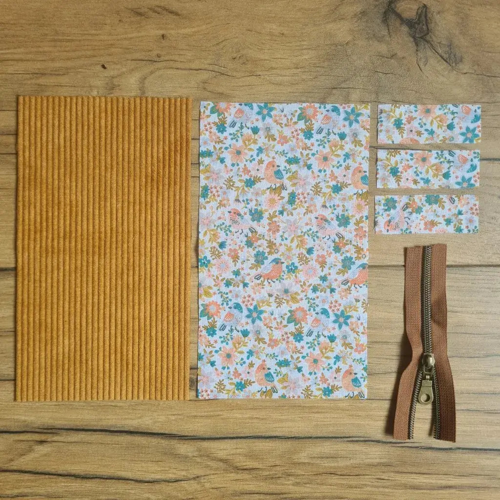 Kit "prêt à coudre" pour une trousse berlingot en velours côtelé au coloris jaune avec une doublure en tissu à motif oiseaux et fleurs. L'ensemble des pièces de tissu, déjà prédécoupées aux bonnes dimensions, sont disposées au sol. A gauche, une pièce en velours côtelé jaune. A droite, quatre pièces en tissu imprimé à motif oiseaux et fleurs ainsi qu'une fermeture éclair marron.