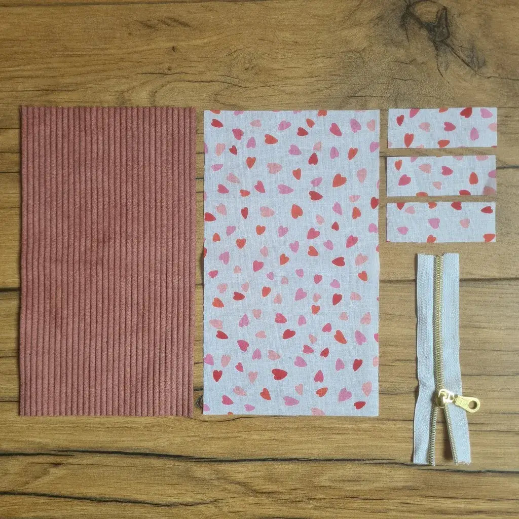 Kit "prêt à coudre" pour une trousse berlingot en velours côtelé au coloris rose avec une doublure en tissu à motif cœurs. L'ensemble des pièces de tissu, déjà prédécoupées aux bonnes dimensions, sont disposées au sol. A gauche, une pièce en velours côtelé rose. A droite, quatre pièces en tissu imprimé à motif cœurs ainsi qu'une fermeture éclair blanche.