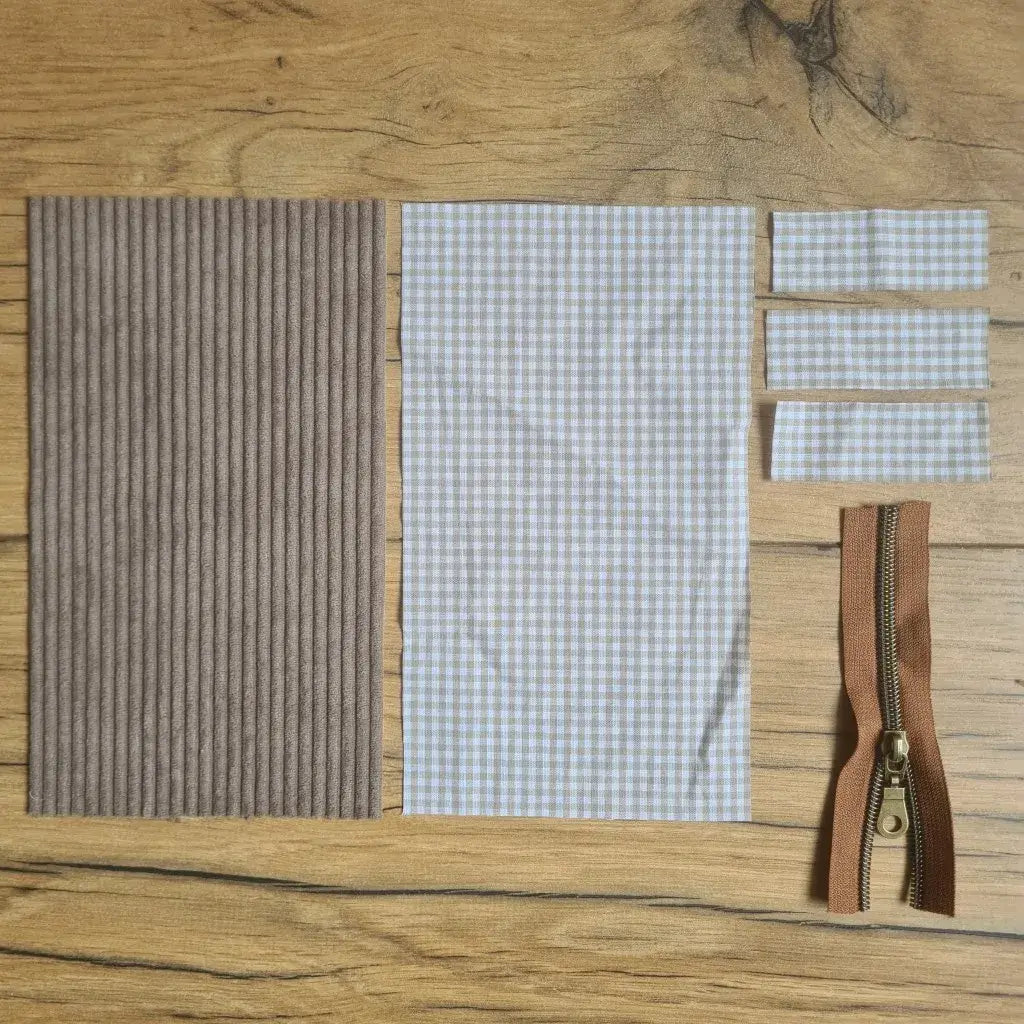 Kit "prêt à coudre" pour une trousse berlingot en velours côtelé au coloris taupe avec une doublure en tissu à motif carreaux beiges. L'ensemble des pièces de tissu, déjà prédécoupées aux bonnes dimensions, sont disposées au sol. A gauche, une pièce en velours côtelé taupe. A droite, quatre pièces en tissu imprimé à motif carreaux beiges ainsi qu'une fermeture éclair marron.