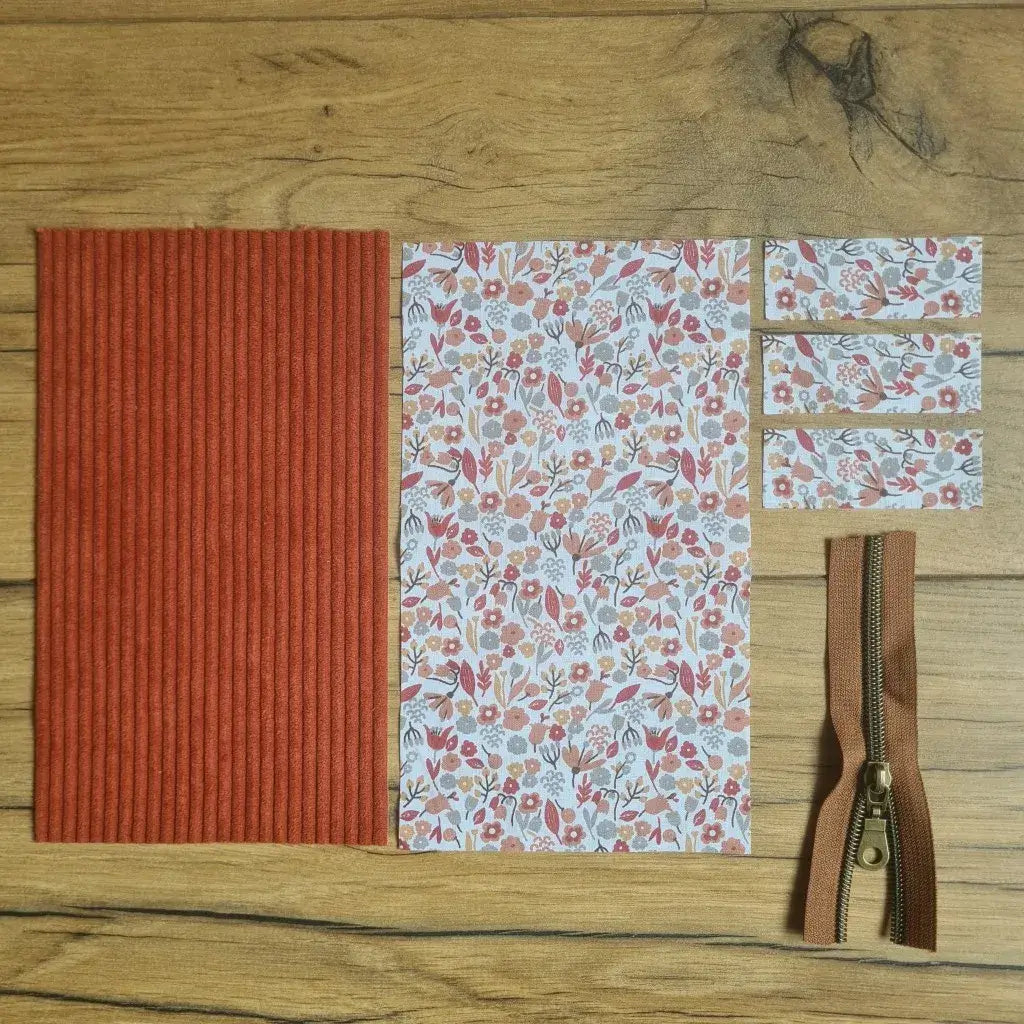 Kit "prêt à coudre" pour une trousse berlingot en velours côtelé au coloris terracotta avec une doublure en tissu à motif liberty. L'ensemble des pièces de tissu, déjà prédécoupées aux bonnes dimensions, sont disposées au sol. A gauche, une pièce en velours côtelé terracotta. A droite, quatre pièces en tissu imprimé à motif liberty ainsi qu'une fermeture éclair marron.