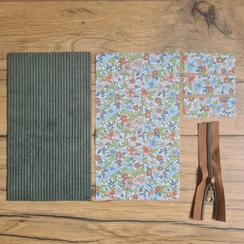 Kit "prêt à coudre" pour une trousse berlingot en velours côtelé au coloris vert avec une doublure en tissu à motif fleurs. L'ensemble des pièces de tissu, déjà prédécoupées aux bonnes dimensions, sont disposées au sol. A gauche, une pièce en velours côtelé vert. A droite, quatre pièces en tissu imprimé à motif fleurs ainsi qu'une fermeture éclair marron.