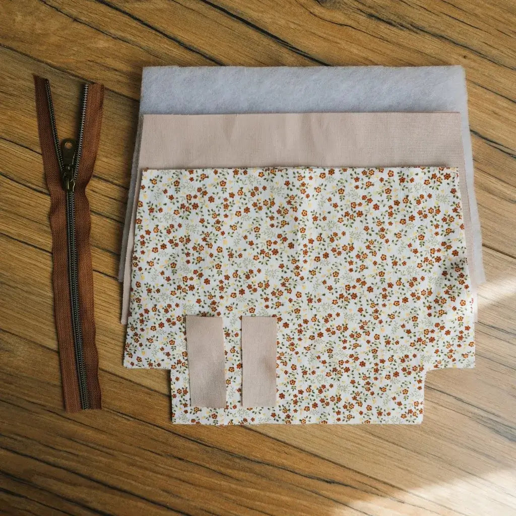 Kit prêt-à-coudre pour réaliser une trousse zippée en tissu imprimé fleuri style prairie, aux petites fleurs orange et jaunes sur fond clair. Le kit contient une fermeture éclair marron avec curseur métallique, des morceaux de tissu découpés, une doublure beige et deux pièces de ouate pour renforcer la tenue. Un projet DIY couture idéal pour créer une trousse fait main pratique et personnalisée.