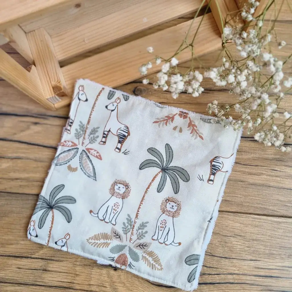 Lingette démaquillante lavable au format maxi 18x18 cm en tissu motif jungle, posée sur un sol en bois. Accessoire réutilisable, zéro déchet et écologique, parfait pour une routine beauté responsable. Imprimé inspiré de la savane avec lions, girafes, zèbres et palmiers, apportant une touche exotique et apaisante. Fabrication artisanale avec couture soignée et texture ultra-douce en coton et éponge de bambou, idéale pour un nettoyage du visage tout en confort et respect de l’environnement.
