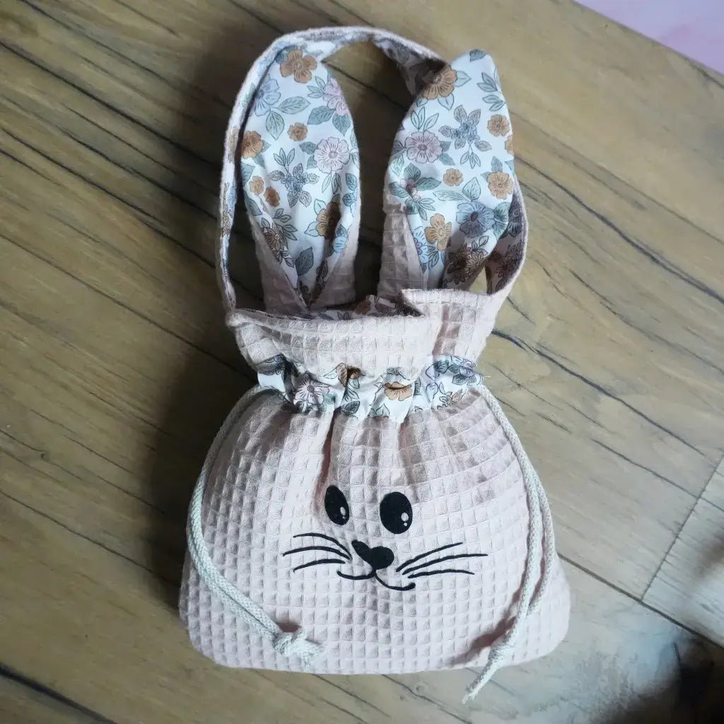 Panier de Pâques lapin en tissu nid d'abeille coloris peau avec oreilles fleuries, posé sur un parquet.