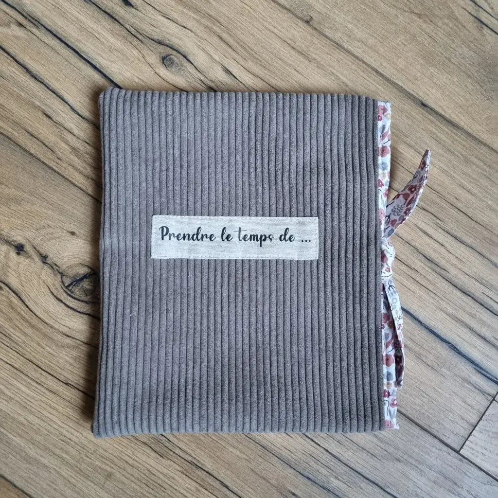 Pochette à livre ou liseuse en velours côtelé taupe avec lien en tissu fleuri et étiquette brodée “Prendre le temps de…”. Accessoire doux et élégant pour protéger un roman, carnet ou liseuse. Fabrication artisanale et finitions soignées. Une idée cadeau originale pour les amateurs de lecture et de créations faites main, mêlant confort, esthétique et praticité.