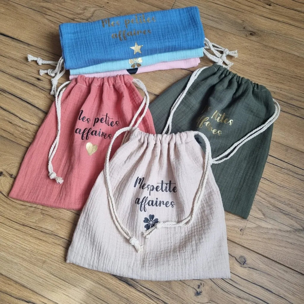 Trois pochons en tissu double gaze de coton sont disposés au sol sur un parquet en chêne. Ces trois pochons sont personnalisés avec l'inscription "Mes petites affaires" floquée en écriture noire ou dorée. Ces pochons se ferment grâce à deux cordelettes en coton blanc. A l'arrière sont pliés et empilés d'autres pochons du même type de coloris divers.