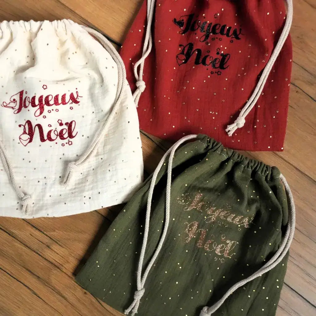 Trois pochons en double gaze décorés du message “Joyeux Noël” en flocage pailleté, présentés dans des teintes festives : blanc, rouge et vert sapin. Chaque sac est orné de petits pois dorés et fermé par un cordon en coton. Ces créations artisanales apportent une touche élégante et naturelle à l’emballage des cadeaux de Noël, alliant douceur du tissu, éclat des paillettes et charme d’un fait main authentique.