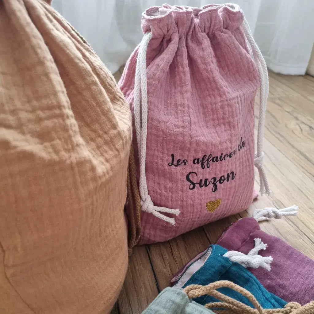 Gros plan sur un sac au format pochon en tissu double gaze de coton de coloris rose sur lequel est floquée une inscription personnalisée "Les affaires de Suzon" en écriture noire avec un petit cœur doré en-dessous. Ce pochon se ferme grâce à deux cordelettes en coton blanc.