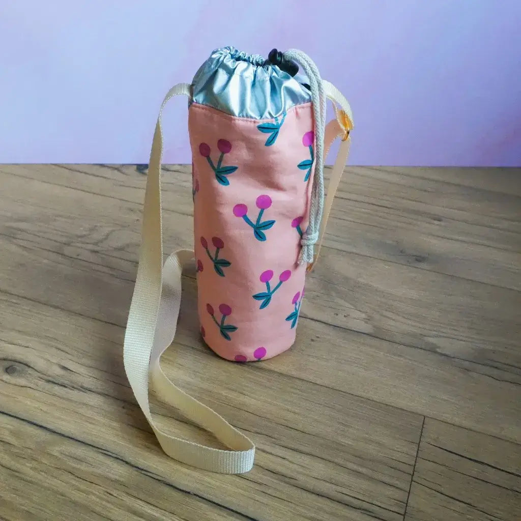Porte-gourde isotherme rose à motif cerises fuchsia avec bandoulière beige et cordon de serrage blanc.