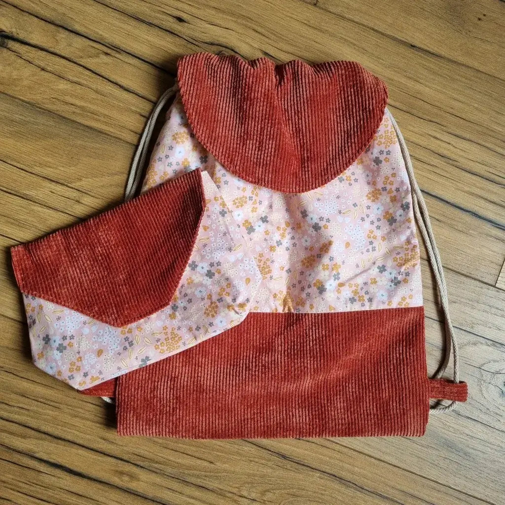 Sac à dos pochon fait main en tissu fleuri rose et velours côtelé terracotta, assorti à une pochette coordonnée avec rabat. Ensemble artisanal idéal pour une rentrée personnalisée, un cadeau unique ou le rangement du quotidien. Fermeture par cordons coulissants, posé sur un sol en bois.