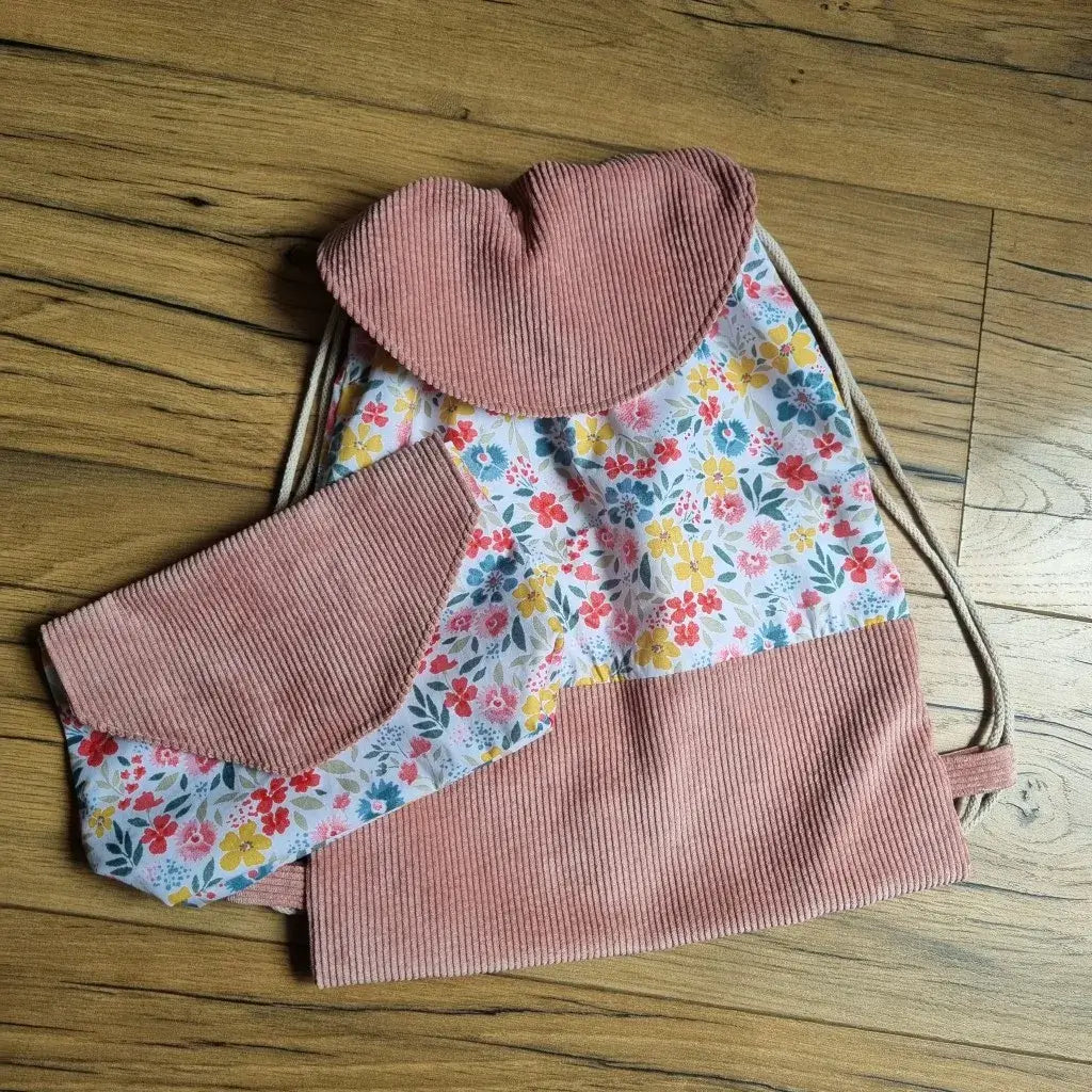 Sac à dos pochon avec rabat assorti et pochette coordonnée, faits main en tissu fleuri multicolore et velours côtelé rose. Le sac se ferme par cordons coulissants beiges et présente un rabat arrondi. Ensemble idéal pour la maternelle, les sorties ou comme cadeau personnalisé. Les finitions soignées et le style bucolique en font un duo à la fois pratique et charmant, mis en valeur sur un fond en bois naturel.