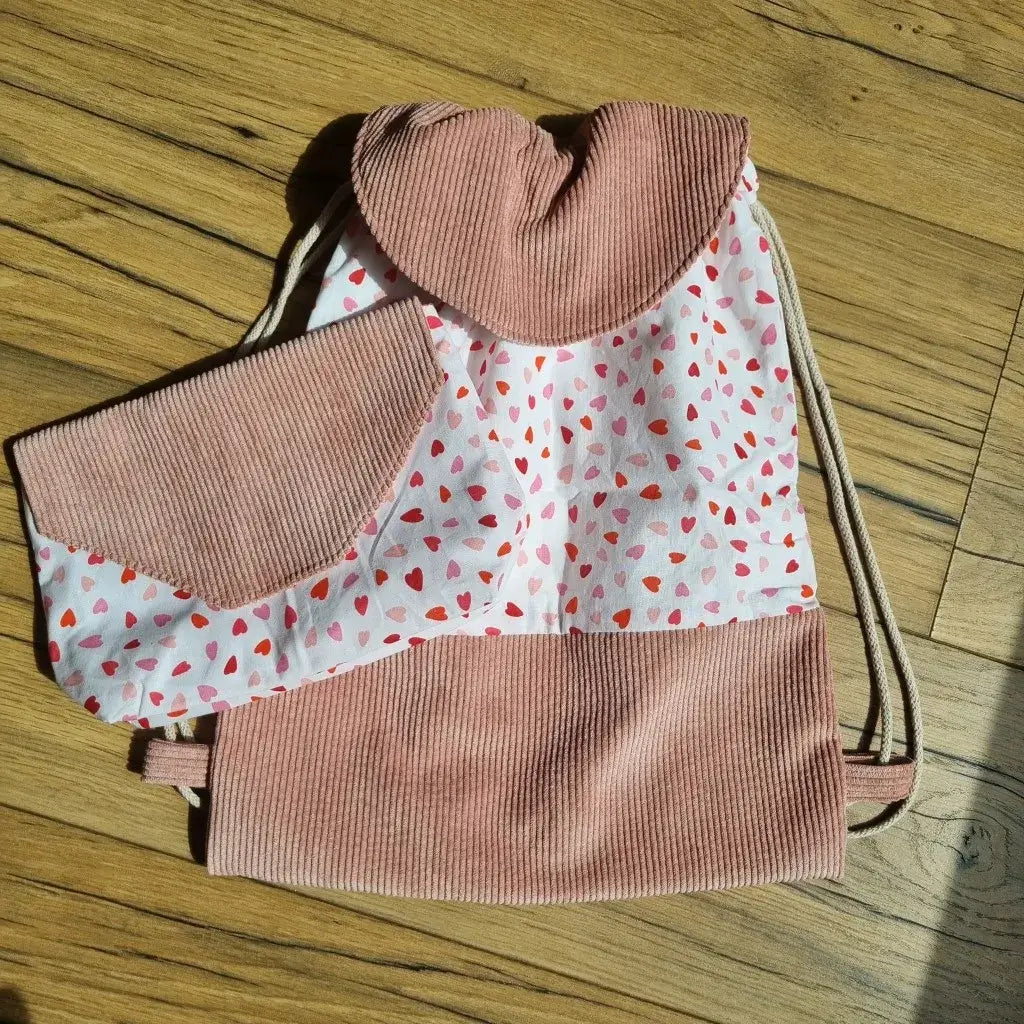 Sac à dos pochon enfant avec rabat en velours côtelé rose, assorti à une pochette coordonnée. L'ensemble est réalisé en tissu à motifs cœurs rouges et roses sur fond blanc, alliant douceur et originalité. Accessoires faits main, parfaits pour la maternelle, les sorties ou comme idée cadeau personnalisée. Posés sur un parquet en bois, ils mettent en valeur un design soigné et une finition artisanale.