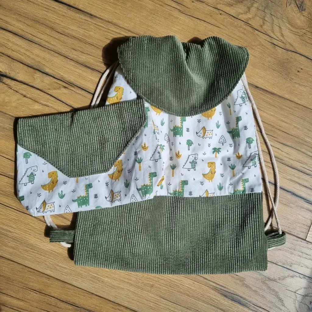 Ensemble sac à dos pochon et pochette assortie pour enfant, confectionnés à la main en velours côtelé vert kaki et tissu imprimé dinosaures colorés sur fond blanc. Le sac dispose d’un rabat arrondi et de cordons beiges pour un port sur le dos. La pochette coordonnée complète l’ensemble, idéal pour la maternelle, la crèche ou les sorties. Accessoires artisanaux ludiques, pratiques et personnalisables, parfaits en cadeau.