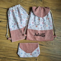 Ensemble personnalisé comprenant deux sacs à dos pochon en tissu imprimé licornes, fleurs et papillons sur fond rose pâle, avec base et rabat en velours côtelé rose, dont un sac avec le prénom Juliette. Pochette assortie posée en bas, idéal pour enfant ou cadeau de rentrée, présenté sur un sol en bois.
