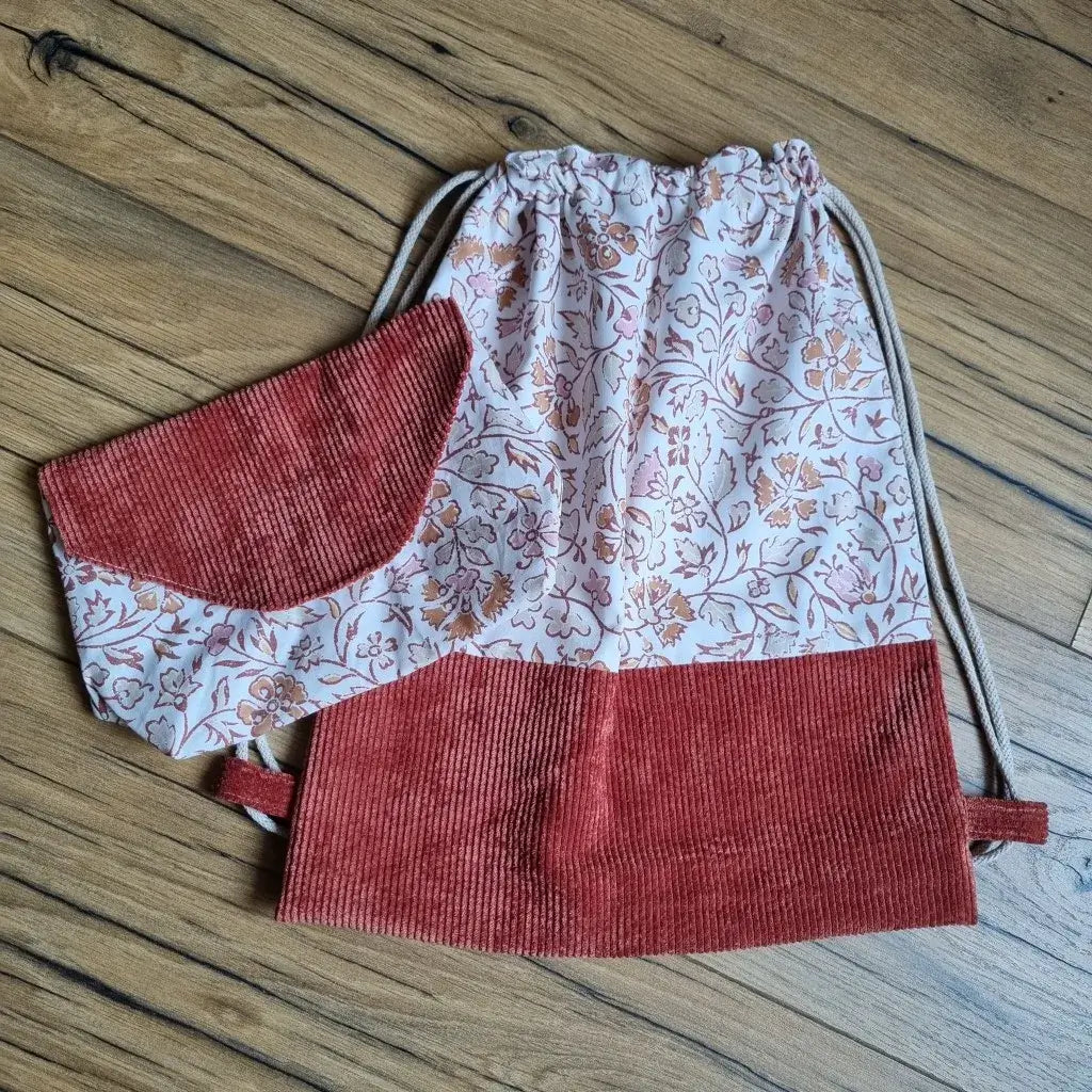 Sac à dos pochon sans rabat en tissu floral oriental rose et terracotta, avec base en velours côtelé terracotta et cordons écrus, accompagné d’une pochette assortie posée sur le sac. Ensemble fait main, idéal pour la maternelle, les loisirs ou comme idée cadeau personnalisé, présenté à plat sur un sol en bois.