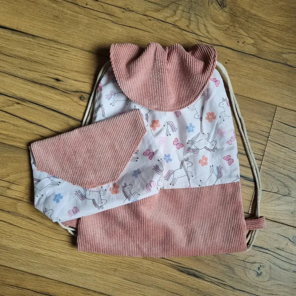 Sac à dos pochon avec rabat et base en velours côtelé rose, en tissu imprimé de licornes, fleurs et papillons sur fond rose pâle, accompagné d’une pochette assortie. Ensemble fait main, idéal pour les enfants, la maternelle ou comme cadeau personnalisé, présenté à plat sur un sol en bois.