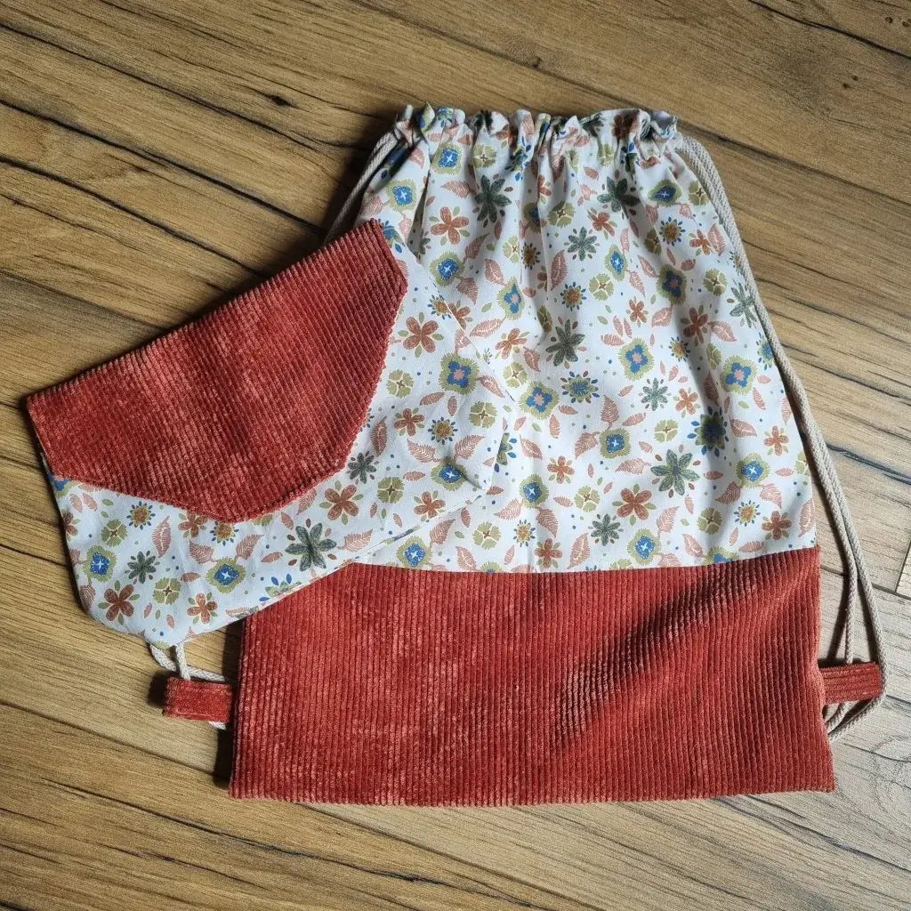 Sac à dos pochon en tissu fait main avec bas en velours côtelé terracotta et haut en coton à motif floral Rozia, accompagné d’une pochette assortie posée par-dessus. Ensemble léger et pratique, idéal pour la maternelle ou les sorties, présenté sur un sol en bois.