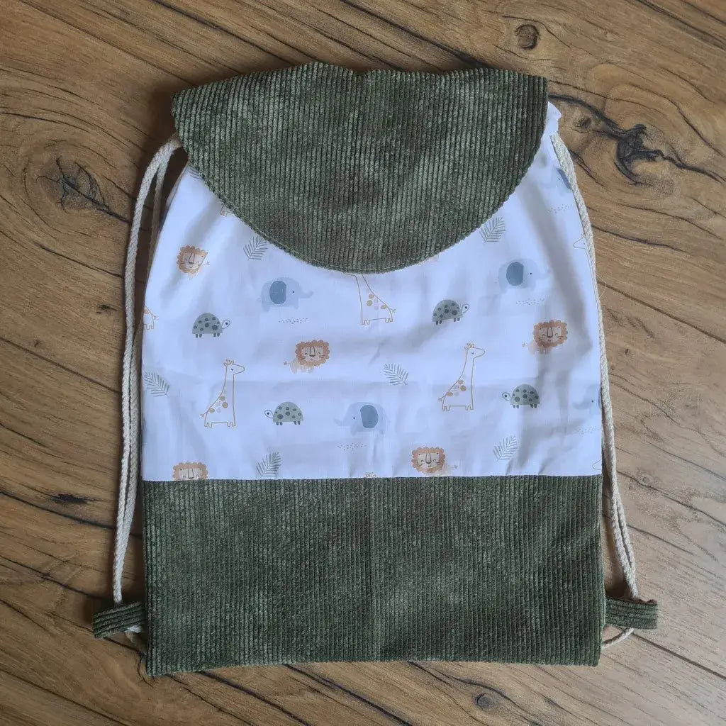 Sac à dos pochon pour enfant avec rabat en velours côtelé vert, tissu imprimé motif savane avec lions, girafes, éléphants et tortues. Fermeture par cordons coulissants, idéal pour la crèche ou les sorties. Accessoire cousu main, pratique et léger, présenté sur un sol en bois. Design ludique et doux, parfait pour un cadeau personnalisé ou une rentrée en maternelle: