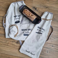 Trois sacs à pain en coton de trois formats différents sont disposés au sol avec un moule à cake et une baguette de pain à l'intérieur. Celui le plus à gauche est au format miche de pain, celui du centre est un sac à cake et celui le plus à droite est au format baguette. Les trois sacs comportent une inscription floquée "mon sac à pain" ou "mon sac à cake" selon l'usage avec un petit logo juste en dessous.