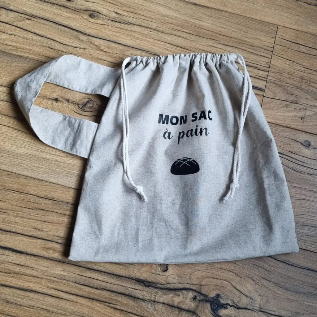 Sac à pain en coton au format miche se fermant par deux petites cordelettes blanches avec une sangle au motif uni du même coloris que l'ensemble et l'inscription "Mon sac à pain" et le logo d'une miche de pain floqués au centre sur la partie supérieure haute.