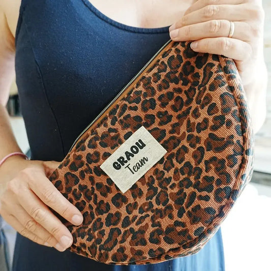 Sac banane demi-lune en tissu imprimé léopard marron et noir, tenu à deux mains par une personne portant une robe bleu marine. Le sac est doté d’une fermeture zippée et orné d’une étiquette cousue sur le devant avec l’inscription “GRAOU Team”. Accessoire pratique et audacieux, parfait pour transporter ses effets personnels tout en affirmant un style sauvage et tendance.