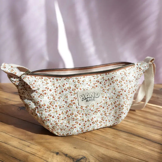 Sac banane demi-lune en tissu crème orné d’un imprimé fleuri miniature dans des tons orange, rouge et vert, posé sur une table en bois éclairée par la lumière naturelle. Équipé d’une bandoulière réglable beige et d’une fermeture zippée, il est agrémenté d’une étiquette “HAND MADE” cousue sur le devant. Accessoire pratique et artisanal, idéal pour transporter l’essentiel avec une touche champêtre et bohème.
