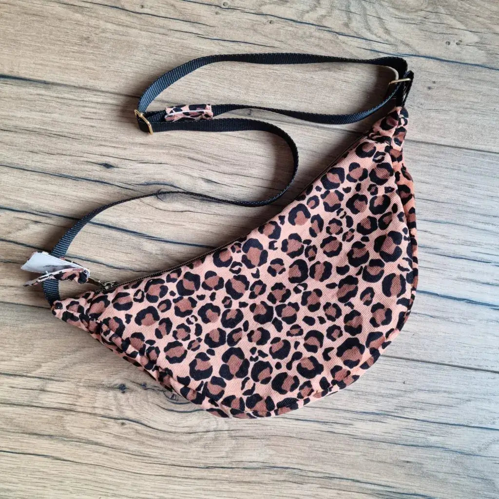 Sac banane demi-lune motif léopard avec sangle noire réglable et zip, posé à plat sur un fond en bois clair.