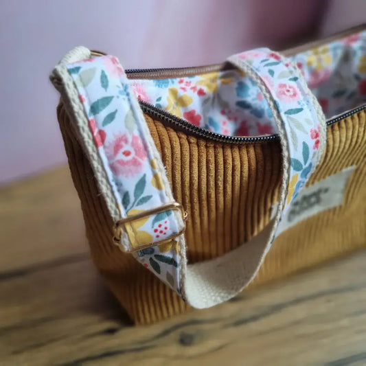 Gros plan sur un sac bandoulière en velours côtelé jaune avec anse réglable en sangle fleurie aux motifs roses, jaunes et verts. La fermeture éclair marron est ouverte, laissant entrevoir la doublure intérieure assortie. L'image met en valeur la qualité des finitions, le tissu texturé et le design floral du sac. Fond en bois et mur rose flouté pour une ambiance douce et naturelle.