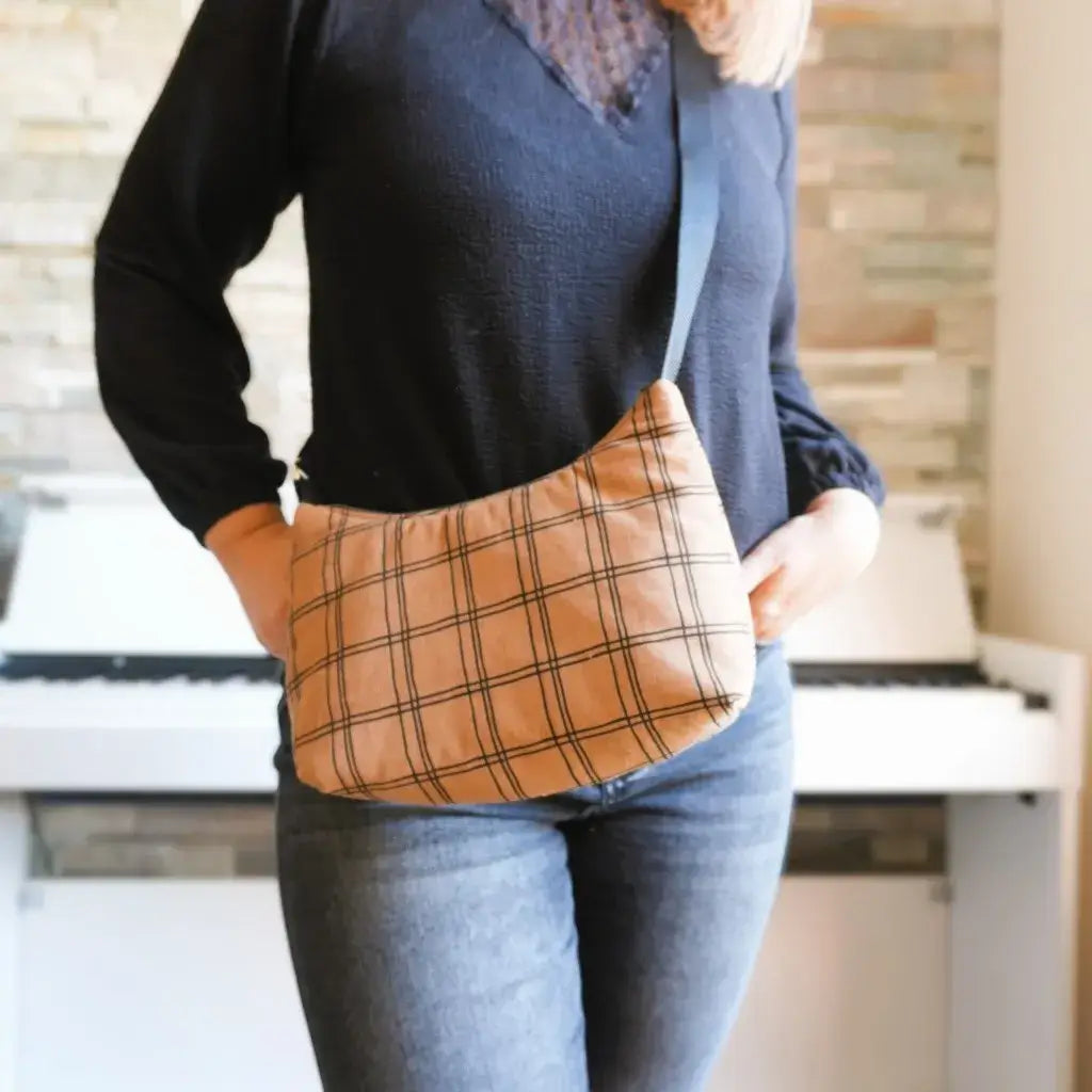 Sac bandoulière en tissu à motif carreaux beige et noir porté en travers du corps par une femme vêtue d’un pull noir et d’un jean. Sac pratique et léger, idéal pour un style décontracté et tendance, parfait pour emporter l’essentiel au quotidien. Photo lifestyle en intérieur sur fond de piano et mur en pierres pour un rendu chaleureux.