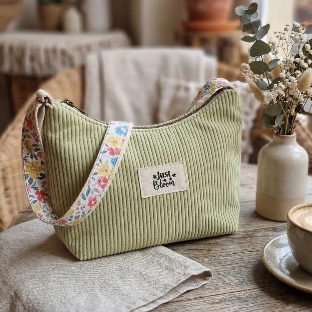 Sac bandoulière en velours côtelé vert anis avec anse fleurie et écusson "Just Bloom" sur une table.