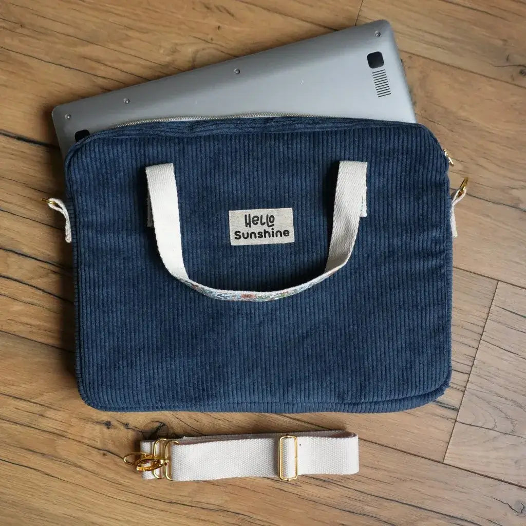 Sacoche pour ordinateur portable en velours côtelé bleu foncé avec poignées en sangle beige et bandoulière amovible, fermeture éclair dorée et étiquette floquée “Hello Sunshine”. Housse de laptop rembourrée, élégante et pratique pour transporter un PC en toute sécurité au travail, à l’école ou en déplacement.