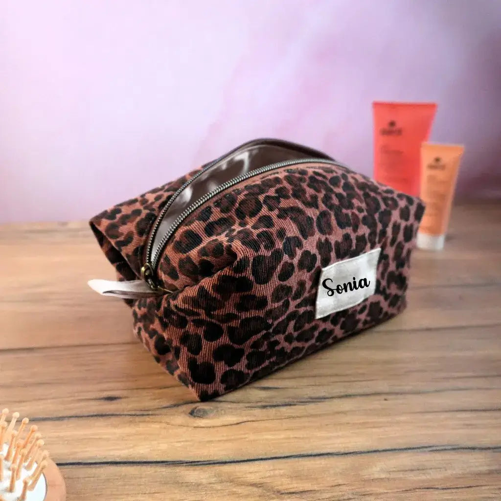 Trousse carrée personnalisée en velours côtelé à motif léopard brun et noir, avec fermeture éclair métallique et étiquette blanche floquée du prénom “Sonia”. Accessoire chic et pratique pour ranger maquillage, bijoux ou produits de toilette. Fabrication artisanale française, design animalier élégant pour un cadeau original et tendance.