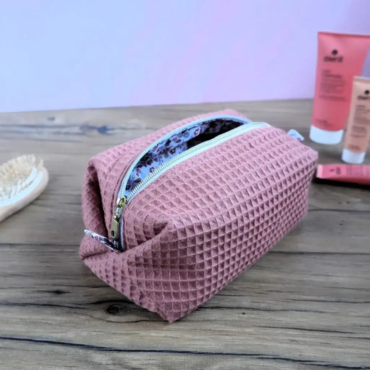 Trousse carrée en tissu nid d’abeille coloris marsala, posée sur une table en bois avec des produits de beauté en arrière-plan. Fermeture éclair dorée et doublure intérieure à motif floral, confection artisanale soignée. Accessoire de rangement élégant et pratique pour maquillage, soins ou voyage. Style moderne et féminin, idéale comme idée cadeau personnalisée ou trousse de bain chic faite main.