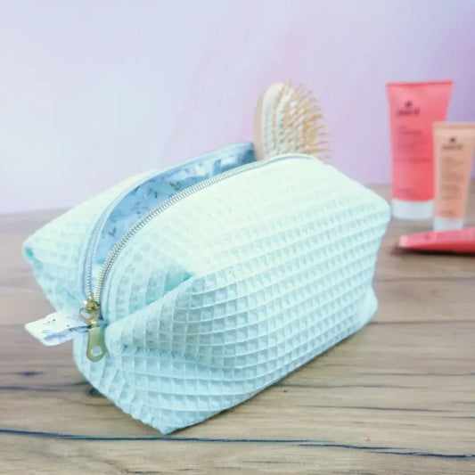 Trousse carrée en tissu nid d’abeille vert menthe, ouverte sur une table en bois avec une brosse et des soins cosmétiques à l’arrière-plan. Fermeture éclair dorée et doublure claire à motif délicat. Accessoire artisanal fait main, léger et pratique pour ranger maquillage, accessoires de beauté ou produits de voyage. Design frais et tendance, parfait comme cadeau personnalisé ou trousse de bain élégante.
